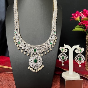 May include: A long, white and green necklace with a large pendant and matching earrings. The necklace is made of white stones and has a green stone in the center of the pendant. The earrings are also made of white stones with a green stone in the center.