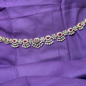 May include: A gold-toned belt with alternating ruby-red and clear gemstone accents. The belt features a floral design with a series of round red stones and smaller clear stones. The belt is displayed on a purple fabric background.