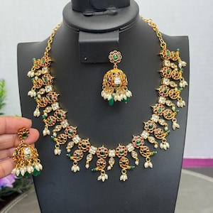 May include: A gold-toned necklace and earring set displayed on a black mannequin. The jewelry features red, green, and clear gemstones, along with small white pearls. The necklace and earrings have a floral design.