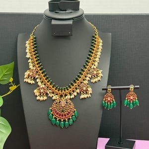 May include: A gold-toned necklace and matching earrings set. The necklace features a central pendant with red and green accents, surrounded by green beads and white pearls. The earrings mirror the design with red and green elements.