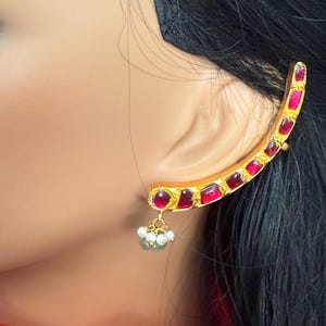 May include: A gold-toned ear cuff with a curved design, adorned with vibrant red gemstones. A cluster of small white beads and a green bead dangle from the bottom. The jewelry piece is worn on the ear.