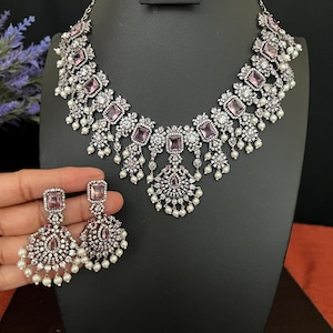 May include: A silver necklace and earrings set with pink and white stones. The necklace has a large pendant with a floral design and the earrings are drop style with a similar design.