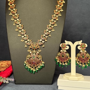 May include: A gold-toned necklace and matching earrings set. The necklace features a pendant with red and green gemstones, and a series of decorative elements. The earrings mirror the pendant design. The set is displayed on a black mannequin.