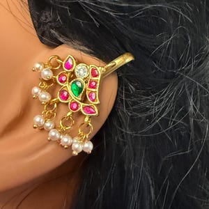 May include: Gold-toned ear cuff with a floral design. The cuff features a central green gemstone, surrounded by pink and clear stones. Small white pearls dangle from the bottom of the cuff, adding a delicate touch. The cuff is designed to clip onto the ear.
