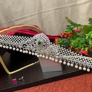 May include: A silver and gold waist belt with a delicate design featuring peacocks and a green stone. The belt is adorned with white pearls and has a matching chain necklace and earrings.