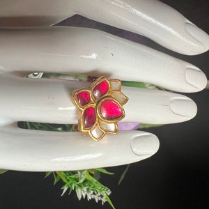 May include: A gold ring with a pink and white flower design. The flower has five petals and is set with red and white gemstones.