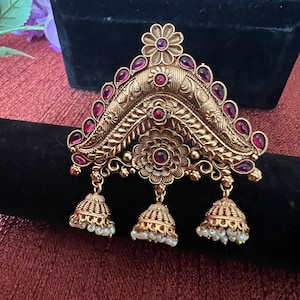 May include: Ornate gold-toned jewelry piece with a floral design and teardrop-shaped red gemstones. The pendant has three bell-shaped dangles with small white beads. The jewelry is displayed on a black surface.