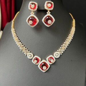 May include: A gold necklace and earrings set with red gemstone accents and clear cubic zirconia stones. The necklace features a V-shaped design with three red gemstones.