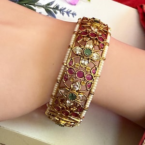 May include: Gold-toned bangle bracelet with intricate floral and geometric designs. The bracelet features red, clear, and green gemstones, and is accented with small white pearls. A beautiful piece of jewelry.