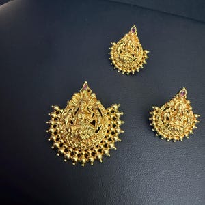 May include: A set of three gold-colored pendants and earrings. The pendants and earrings feature a detailed design of a seated figure, possibly a deity, with a small red gem at the top. The jewelry has a beaded border.