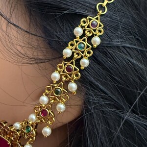 May include: Gold-toned hair jewelry with star-shaped accents, red, green, and white gemstones, and dangling pearls. The jewelry is worn near black hair. The design is ornate and detailed, suitable for special occasions.