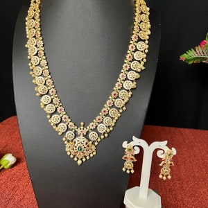 May include: A gold necklace and earrings set with white and green stones, and red accents. The necklace has a pendant with a pearl drop.