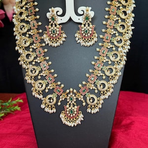 May include: A gold-toned necklace and earring set with intricate detailing. The necklace features a series of connected circular and square elements, adorned with red, green, and white accents. The matching earrings have a similar design, with a teardrop shape and dangling pearls.