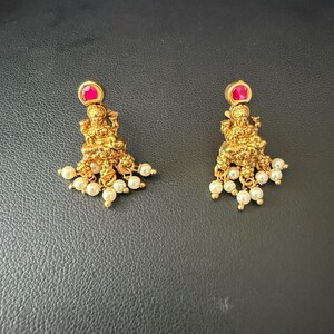 May include: A pair of gold earrings with a small red stone and pearl accents. The earrings feature a detailed design of a seated figure.