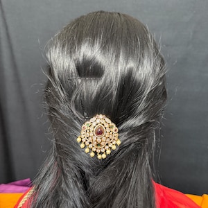 May include: A gold hair clip with a large red gemstone surrounded by smaller white and green gemstones. The clip has a dangling pearl accent.