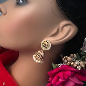 May include: Gold earrings with a peacock design and red and white stones. The earrings are dangling and have a pearl drop.