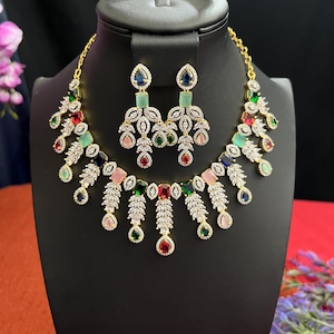 May include: A gold necklace and earrings set with a cascading design of clear and colorful stones. The necklace features a row of square-shaped stones in shades of pink, red, green, blue, and light blue. The earrings have a similar design with a teardrop-shaped stone at the bottom.