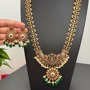 May include: A gold-toned necklace and matching earrings set. The necklace features a detailed peacock design with red, green, and white gemstones. The earrings have a similar design with green beads.
