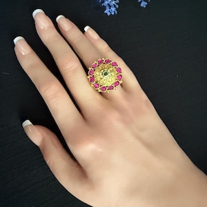 May include: A gold-toned ring with a circular design. The ring features a central green stone surrounded by a pattern of pink gemstones. The ring is worn on a hand with manicured nails.