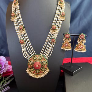 May include: An ornate necklace and earring set. The necklace features multiple strands of white beads with gold accents and red and green floral designs. The large pendant has a red center and is surrounded by green and gold filigree. The earrings mirror the design.