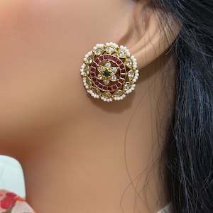 May include: Gold-toned stud earrings with a circular design. The earrings feature a central flower design with a green stone, surrounded by red stones, clear crystals, and white pearls. The earrings are set against a skin tone background.