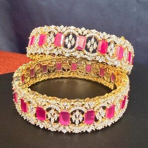 May include: Two gold-toned bangles with a repeating pattern of rectangular pink gemstones and clear, sparkling accents. The bangles are displayed on a dark surface, showcasing their intricate design and vibrant colors.