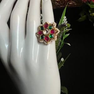 May include: A gold ring with a flower design. The flower has red, green, and white gemstones. The ring is on a white mannequin hand.