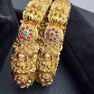 May include: Two ornate gold bangles. Each bangle features intricate carvings of figures, floral designs, and circular accents with red and green gemstones. The bangles are displayed against a dark background, highlighting their detailed craftsmanship and rich color.