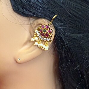 May include: A gold-toned earring with a floral design, featuring red and green gemstones, and small white pearls. The earring is worn on an ear, with black hair in the background. The earring has a hook for attachment.