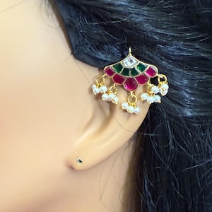 May include: A gold-toned earring with a fan-shaped design, featuring red, green, and white enamel accents. Small white beads dangle from the bottom. The earring is worn on a person's ear.