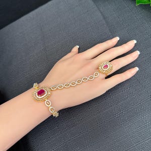 May include: A gold bracelet with a red stone and a matching ring. The bracelet has a chain that connects to the ring, allowing it to be worn on the finger.