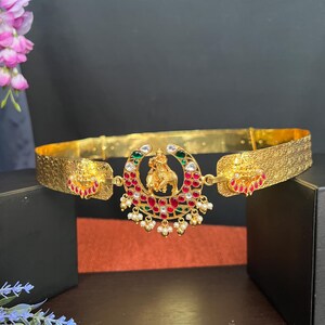 May include: A gold waist belt with a decorative center piece featuring an elephant and red, green, and white gemstones. The belt is made of a textured metal and has a simple design.