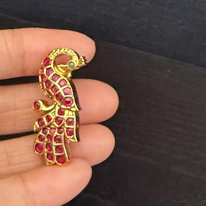 May include: A gold-toned peacock brooch with red and green gemstones. The peacock is depicted in a standing position with its tail feathers spread out.