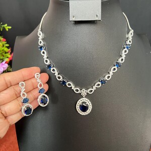 May include: A silver necklace and earrings set with a blue gemstone pendant and matching earrings. The necklace features a delicate chain with small blue gemstones and a larger oval-shaped pendant with a blue gemstone surrounded by smaller white gemstones. The earrings are drop earrings with a blue gemstone and smaller white gemstones.