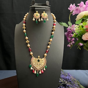 May include: A gold-toned necklace and earring set displayed on a black mannequin. The necklace features a pendant with a central green stone, surrounded by white and red accents. The earrings mirror the pendant design. The necklace has alternating red, green, and gold beads.