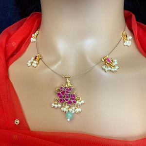 May include: A gold necklace with a pink and gold pendant featuring a floral design. The pendant is adorned with small white pearls and a green stone. The necklace also has three smaller pendants with pink stones and white pearls.