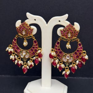 May include: Gold-toned, ornate earrings with a crescent moon design. The earrings feature a floral top, a ruby-colored stone-filled crescent, and dangling red and white beads. The earrings are displayed on a white stand against a black background.