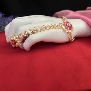 May include: A gold hand and finger ring with a pink flower design and clear stones.