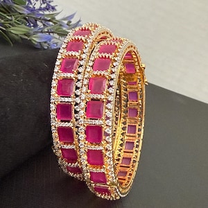 May include: Two gold-colored bangles with a repeating pattern of square, magenta-colored stones. The stones are framed by small, clear crystals. The bangles are displayed on a dark surface, with a sprig of purple flowers in the background.