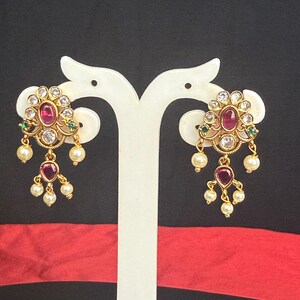 May include: Gold-toned earrings with a floral design, featuring a central ruby-colored stone surrounded by clear crystals. Small green stones and dangling faux pearls add detail. The earrings are displayed on a white stand against a black and red background.