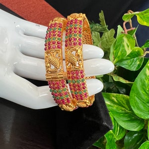 May include: Two gold bangles with intricate designs and red and green gemstones. The bangles are on a white surface.