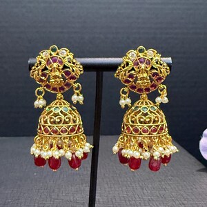 May include: A pair of gold-toned jhumka earrings featuring a circular top with a central deity figure, accented with red and green stones. The bell-shaped base is decorated with intricate filigree, red stones, and dangling white and red beads.
