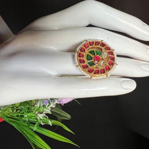 May include: A gold-toned ring with a circular design. The ring features a central design with green and clear stones, surrounded by red stones and small white pearls. The ring is set on a white hand.