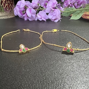 May include: Two gold anklets with a floral design. Each anklet features a central cluster of gemstones: a white, a green, and two red stones. The anklets are displayed on a dark surface, with purple flowers in the background.