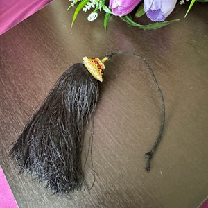 May include: A black tassel with a gold and red jeweled top. The tassel is attached to a black cord.