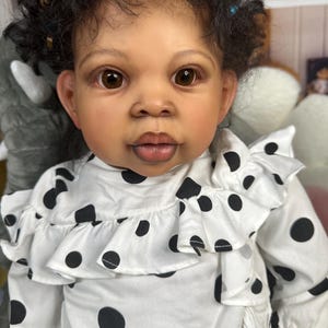 Może przedstawiać: Zbliżenie na lalkę niemowlęcą z brązowymi oczami i ciemnymi, kręconymi włosami, ubraną w białą sukienkę w czarne kropki z falbanką.