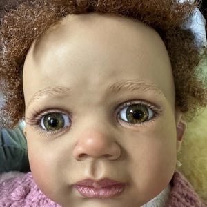May include: A close-up of a doll's face with brown curly hair, brown eyes, and a pink knitted sweater.