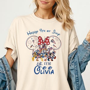 Pode incluir: Camiseta cor marfim com um design patri&oacute;tico com orelhas do Mickey Mouse, um la&ccedil;o vermelho e o texto "Happy 4th of July". O design inclui personagens de desenhos animados e o nome "Olivia" em azul.