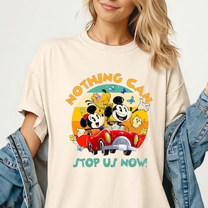 May include: Ivory-colored t-shirt featuring a colorful graphic of Mickey and Minnie Mouse in a red car, with the text "NOTHING CAN STOP US NOW!". The design includes a yellow sun, butterflies, and flowers.
