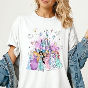 K&ouml;nnte beinhalten: Wei&szlig;es T-Shirt mit einer farbenfrohen Illustration von Prinzessinnen vor einem Schloss. Das Design umfasst rosa, blaue und lila Akzente mit Feuerwerk und Luftballons. Das Shirt ist mit "Comfort Colors White" gekennzeichnet.